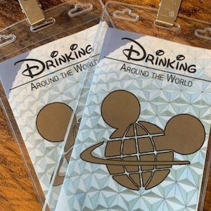 May include: Two blue and gold "Drinking Around The World" event passes. The passes feature a gold Mickey Mouse head design over a globe with the words "Drinking Around The World" in white text.