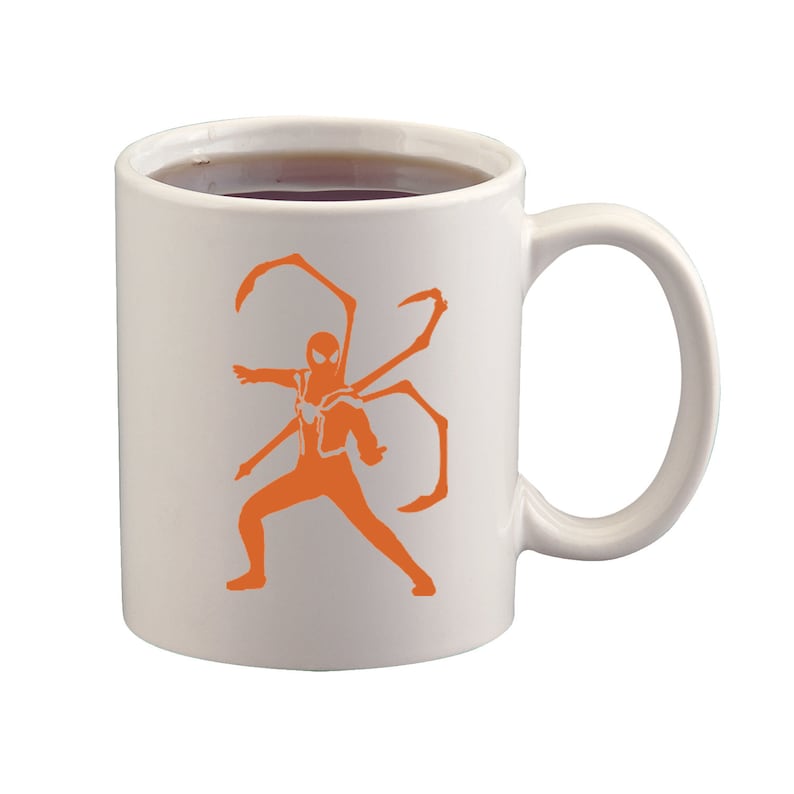 May include: White ceramic mug with a handle, filled with a dark liquid. The mug features an orange silhouette of a superhero with multiple mechanical arms. The design is centered on the mug's surface.