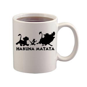 May include: White ceramic mug with a black silhouette design featuring characters from The Lion King, including a lion cub, meerkat, warthog, and the phrase "HAKUNA MATATA". The mug is filled with a dark liquid.