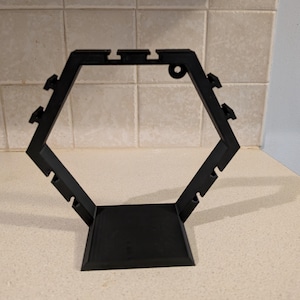 May include: A black, hexagonal display stand with a flat base and a series of notches and holes. The stand is made of a solid material and is designed to hold items. The stand is approximately 8 inches tall.
