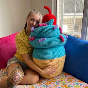 Può includere: Un grande giocattolo di peluche a forma di cono gelato all'uncinetto con glassa blu, gialla e rosa e una ciliegia rossa in cima. Una persona tiene il giocattolo di fronte a sé.