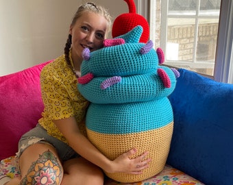 May include: A large crocheted ice cream cone shaped plush toy with blue, yellow, and pink frosting and a red cherry on top. A person is holding the toy in front of them.