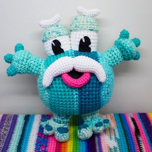 May include: A crocheted blue and green monster with a white mustache and pink tongue. The monster has two large eyes and two small horns on its head. It is standing on a rainbow striped surface.