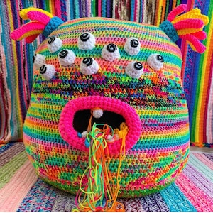 Puede incluir: Un juguete monstruo de crochet colorido con una gran boca abierta y múltiples ojos. El monstruo está hecho con una variedad de colores brillantes, incluyendo rosa, azul, verde, amarillo y naranja. El juguete está hecho de hilo y tiene una superficie texturizada.