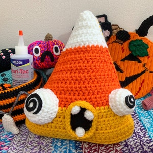 Puede incluir: Un peluche de maíz caramelo tejido a crochet con ojos saltones y una sonrisa de dientes. El peluche es naranja, amarillo y blanco y está sentado sobre una manta de rayas de colores.
