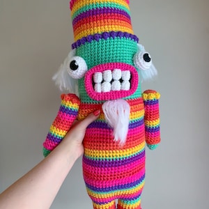 May include: A colorful crocheted toy with a rainbow striped body, green head, white teeth, and large black eyes. The toy has a white beard and a rainbow striped hat.