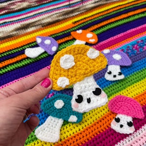 May include: A collection of crocheted mushrooms in various colors, including yellow, purple, teal, pink, and white. The mushrooms have white spots and cute faces. The mushrooms are displayed on a colorful striped crocheted blanket.