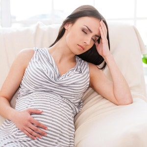 May include: A pregnant woman sits on a white sofa, holding her stomach and looking distressed. She is wearing a striped grey and white dress and has her hand on her forehead.