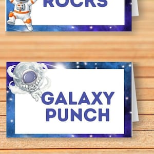 May include: Four rectangular folded paper food labels with a dark blue galaxy border. The labels feature space-themed illustrations of an astronaut, planets, a rocket, and text reading SPACE ROCKS, LUNAR COOKIES, GALAXY PUNCH, and ALIEN CRITTERS.