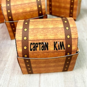 May include: Printable pirate treasure chest party favor boxes. The boxes are brown with gold accents and a brown strap. The box in the foreground has the name "Captain Kim" written on it.