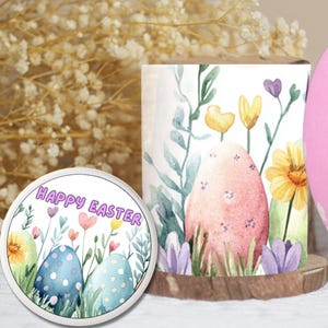 Op de afbeelding: Een Paas-thema display met een roze ei met witte bloemaccenten, een cilindrische kaars met een ei- en bloemontwerp, en een ronde coaster met de tekst "HAPPY EASTER" en geschilderde eieren. De scène wordt geplaatst tegen een achtergrond van gedroogde bloemen.