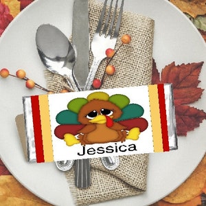May include: Thanksgiving place setting with a white plate, silverware, and a burlap napkin. A candy bar with a turkey graphic and the name "Jessica" is placed on the napkin. The setting is surrounded by colorful autumn leaves.