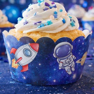 Puede incluir: Primer plano de un cupcake con glaseado blanco y chispas de colores. El cupcake está en un envoltorio azul decorado con cohetes, astronautas y planetas. Otros cupcakes están en el fondo, con un fondo azul y las palabras "HAPPY BIRTHDAY".