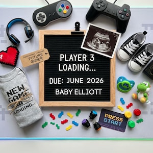 May include: A baby announcement with a gaming theme. A black letter board displays "PLAYER 3 LOADING... DUE: JUNE 2026 BABY ELLIOTT." The image includes a baby scan, small trainers, a babygrow, game controllers, and a Yoshi toy.