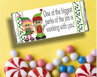 May include: A white chocolate bar wrapper with a festive design featuring two elves holding tools. The text on the wrapper reads "One of the biggest perks of the job is working with you!"