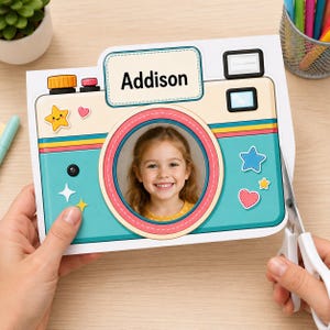 May include: A rectangular card shaped like a camera with a teal body, yellow and pink accents, and a central circular cutout displaying a photograph of a smiling child. The card features the name Addison in black text and various star and heart stickers.