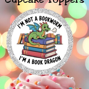 Pode incluir: Um cupcake coberto com glacê rosa e granulado colorido. Um topper circular com o texto "I'M NOT A BOOKWORM I'M A BOOK DRAGON" e um dragão de desenho animado em livros é colocado em cima. As palavras "Book Lover Cupcake Toppers" estão no topo.