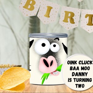 May include: A cylindrical container with a cartoon cow face design. The cow has large eyes, a pink nose, and is eating grass. A round label reads "OINK CLUCK BAA MOO DANNY IS TURNING TWO". A banner spells "BIRTH" in gold letters.