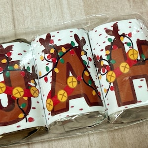 May include: Three individually wrapped chocolate bars with festive reindeer designs. Each bar features a different letter, spelling out the name "JAH". The reindeer are decorated with red and green lights and gold bells.