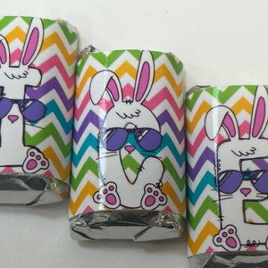 May include: Three chocolate bar wrappers with a colorful chevron pattern. Each wrapper features a cartoon bunny wearing sunglasses. The bunnies are white with pink ears and paws.