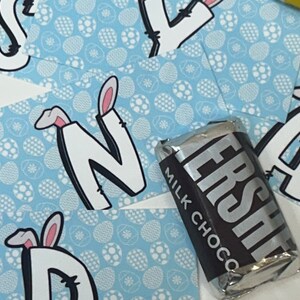 May include: Four blue cards with white bunny ears and the letters N, A, D, and S on them. The cards have a white and blue egg pattern. A silver chocolate bar with the text "Milk Choco" and "ERSHEY'S" is on top of the cards.