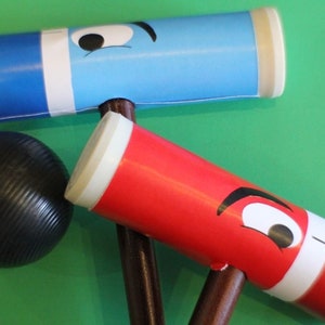 May include: Two wooden croquet mallets with painted faces. One mallet is blue with a white stripe and the other is red with a white stripe. A black croquet ball is in the foreground.