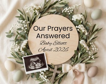 May include: A circular wooden announcement with the text "Our Prayers Answered Baby Elliott April 2026", surrounded by a floral wreath. Includes an ultrasound image, decorative eggs, a wooden cross, and a ribbon.
