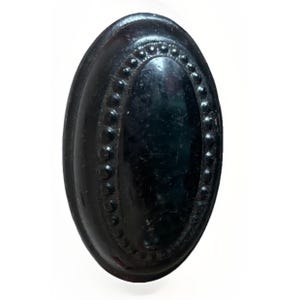 May include: A dark brown oval-shaped button featuring a smooth, glossy center panel surrounded by a raised, textured border with a repeating beaded or dotted pattern along the inner edge of the rim, creating a distinct decorative frame.