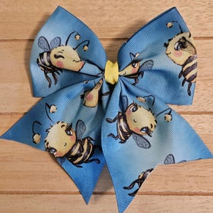 May include: A large, blue hair bow with a repeating pattern of cartoon bees. The bees are yellow and black with smiling faces. The center of the bow is yellow.