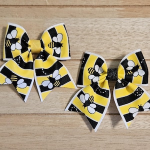 May include: Two yellow, black, and white bee-themed hair bows. The bows feature a striped pattern with bee illustrations. The bows are made of ribbon and are set against a wooden background.