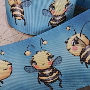 Puede incluir: Un rollo de cinta azul con un patrón repetido de abejas de dibujos animados. Las abejas tienen cuerpos amarillos con rayas negras, ojos azules y mejillas rosadas. La cinta está hecha de un material texturizado.