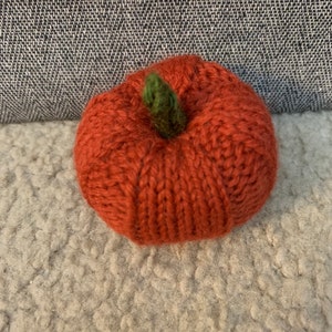 May include: A small, knitted pumpkin with a green stem. The pumpkin is orange and has a textured surface.
