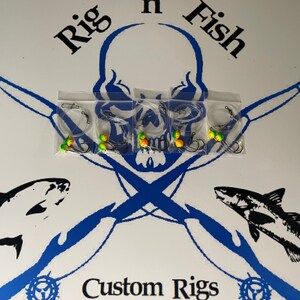 May include: Five clear plastic bags containing fishing rigs with green and yellow beads. The bags are arranged in a row on a white surface with a blue and white skull and crossbones design. The text "Rig n Fish" is above the skull and crossbones and the text "Custom Rigs" is below the skull and crossbones.