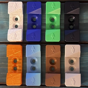 May include: A collection of twelve rectangular, multi-colored, 3D-printed objects. Each object has a fish-shaped design and two circular holes. Colors include white, blue, green, black, orange, light blue, brown, and off-white. The objects are arranged on a blue wooden surface.