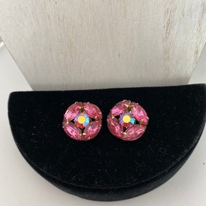 May include: A pair of vintage pink rhinestone earrings. Each earring features a circular design with pink, oval-shaped rhinestones surrounding a central, iridescent stone. The earrings are set on a gold-toned metal base and rest on a black velvet surface.