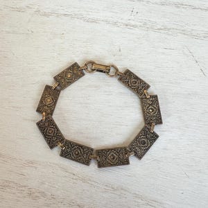 May include: An antique gold-toned bracelet with rectangular links. Each link features an ornate, geometric pattern. The bracelet is connected with a simple clasp. The jewelry is displayed on a white, textured surface.