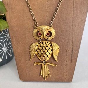 Puede incluir: Un collar de tono dorado con un colgante de búho con ojos de pedrería roja. El búho está detallado con un cuerpo texturizado y una rama con una cola.