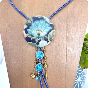 May include: A blue and green floral patterned fabric circle pendant necklace with blue crystal accents and a blue braided cord.