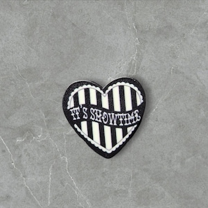 May include: Black and white striped heart-shaped patch with the text "It's Showtime" in white.