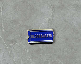 May include: A rectangular enamel pin featuring a deep blue background with a gold-colored border and the word BLOCKBUSTER embossed in gold lettering across the center, designed to resemble a classic movie rental ticket stub.