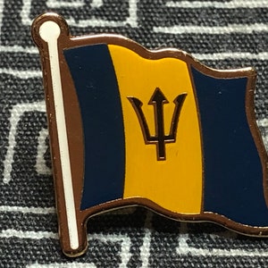 May include: A gold-colored pin with a blue and yellow flag of Barbados. The flag features a gold trident on a yellow field.