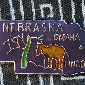 May include: Purple enamel pin shaped like the state of Nebraska with the words "NEBRASKA", "OMAHA", and "LINCOLN" in gold lettering. A green tree and a brown horse are depicted on the pin.