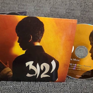 May include: A CD case with a yellow and orange background featuring a silhouette of a person with the text "3121" in white. The CD is visible in the background.