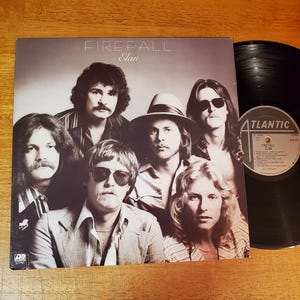 May include: Album cover for Firefall's "Elan" album. The cover features a group of seven men with long hair and sunglasses. The album is next to a black vinyl record with the Atlantic Records logo.