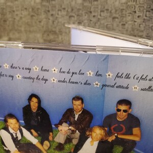 May include: A CD case with a blue background and a group of five people. The text on the case reads "better now", "there's a way", "home", "how do you love", "him", "feels like (it feels alright)", "perfect to stay", "counting the days", "under heaven's skies", "general attitude", and "satellite".