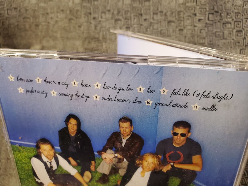 May include: A CD case with a blue background and a group of five people. The text on the case reads "better now", "there's a way", "home", "how do you love", "him", "feels like (it feels alright)", "perfect to stay", "counting the days", "under heaven's skies", "general attitude", and "satellite".
