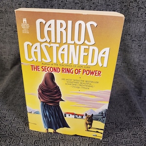 May include: A yellow paperback book with the title "The Second Ring of Power" by Carlos Castaneda. The book cover features a woman in a brown shawl and a dog.