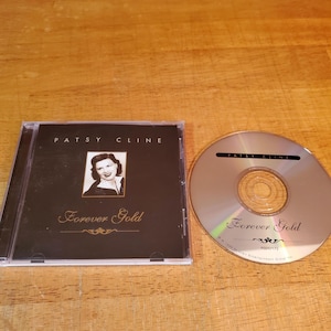 May include: A Patsy Cline "Forever Gold" CD album. The CD case is black with a photo of Patsy Cline and the album title in gold lettering. The CD is silver with the album title and artist's name.