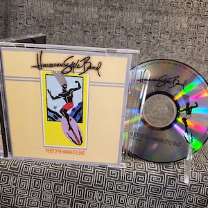 May include: A CD case and disc featuring "The Best of the Hawaiian Style Band." The cover art displays a surfer illustration in a yellow and blue frame. The disc itself reflects the same image. The text "Hawaiian Style Band" is visible.