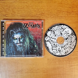 May include: A Rob Zombie Hellbilly Deluxe music CD in a plastic jewel case with a skull-patterned spine and a disc featuring a black and white illustration of various monstrous faces. The cover art displays a portrait with glowing yellow eyes.